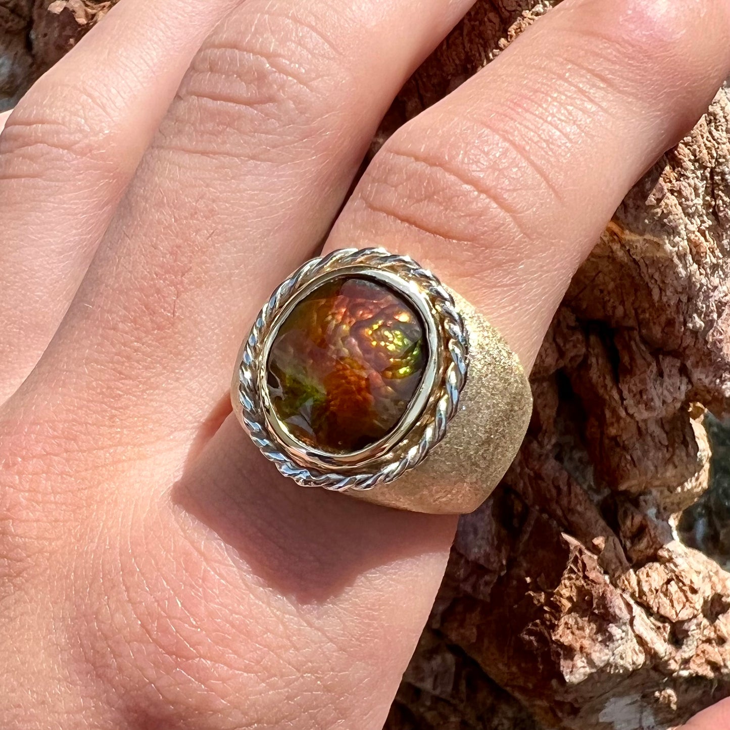 A men's matte finish textured gold fire agate ring.  There is a gold rope bezel around the oval cabochon cut stone.