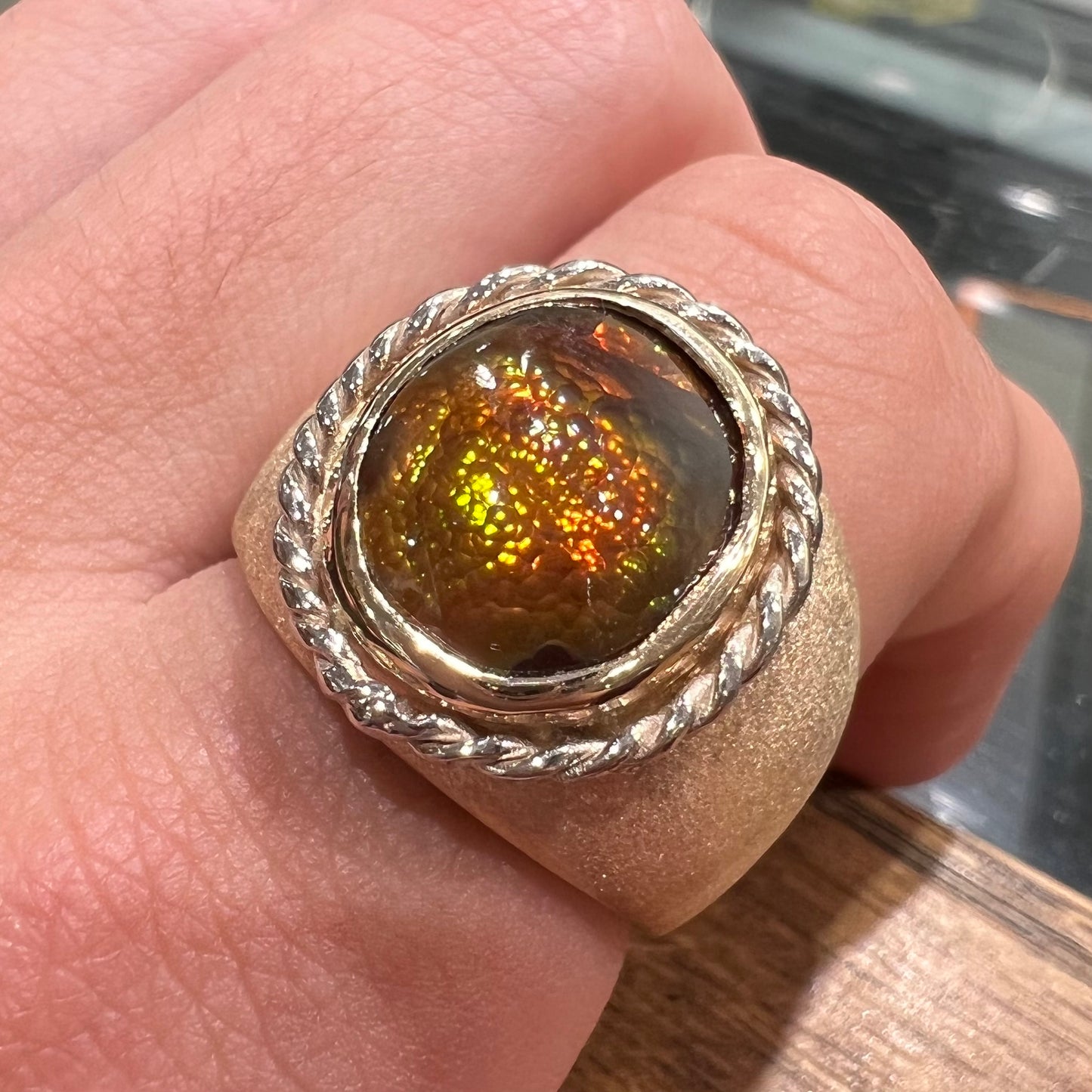 A men's matte finish textured gold fire agate ring.  There is a gold rope bezel around the oval cabochon cut stone.