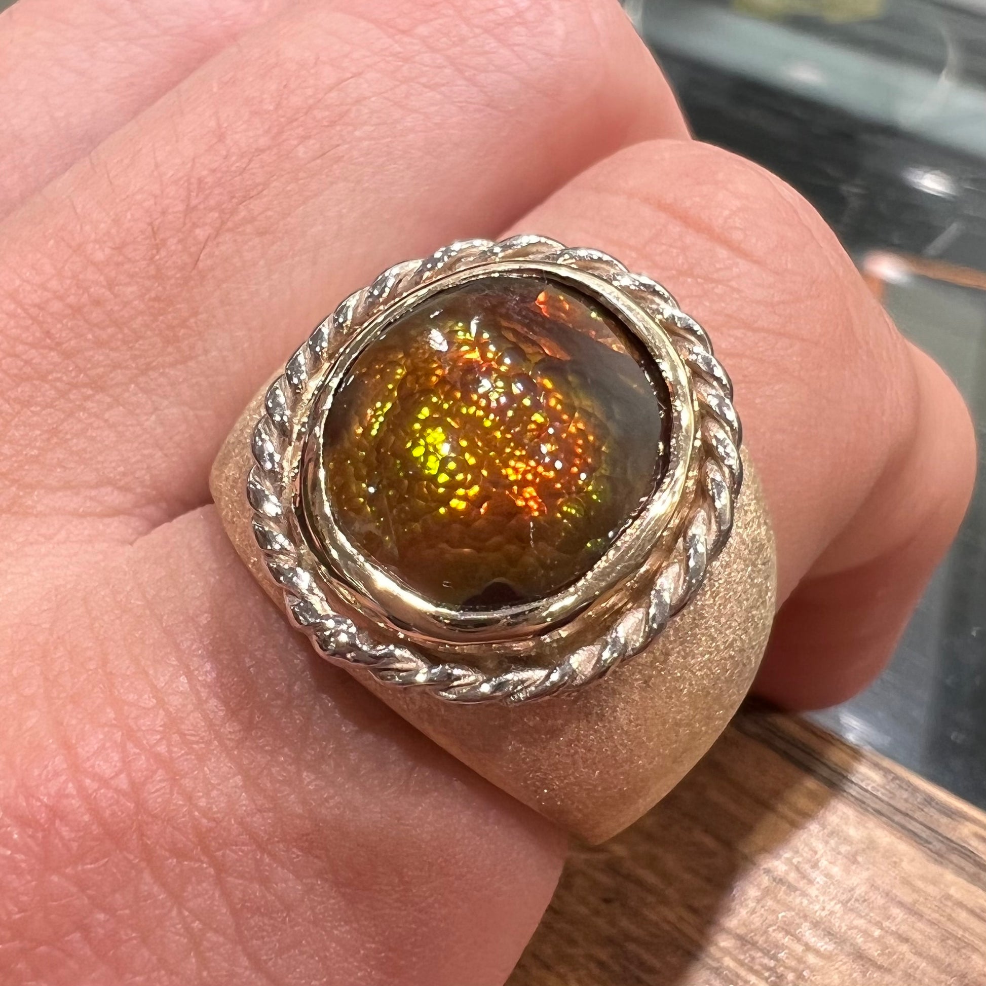 A men's matte finish textured gold fire agate ring.  There is a gold rope bezel around the oval cabochon cut stone.
