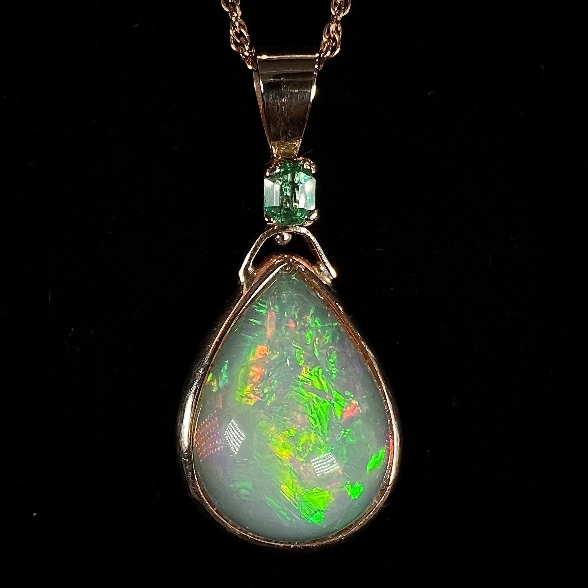 A yellow gold bezel set Ethiopian fire opal drop pendant.  There is an emerald set in the bail.