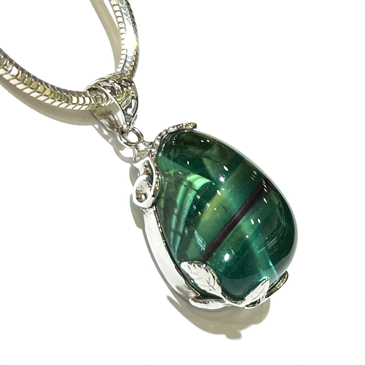 A solitaire gemstone pendant featuring a cabochon-cut pear shape clear green fluorite stone with purple banding on a thick sterling silver snake chain.  The gem is held by prongs shaped like leaves and vines.