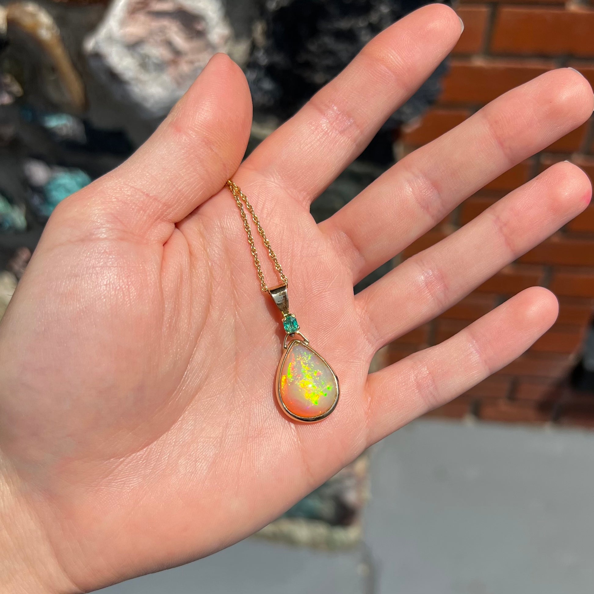 A yellow gold bezel set Ethiopian fire opal drop pendant.  There is an emerald set in the bail.