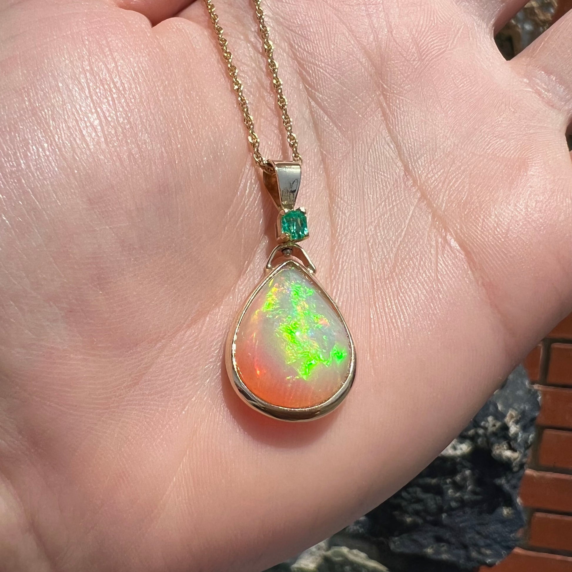 A yellow gold bezel set Ethiopian fire opal drop pendant.  There is an emerald set in the bail.