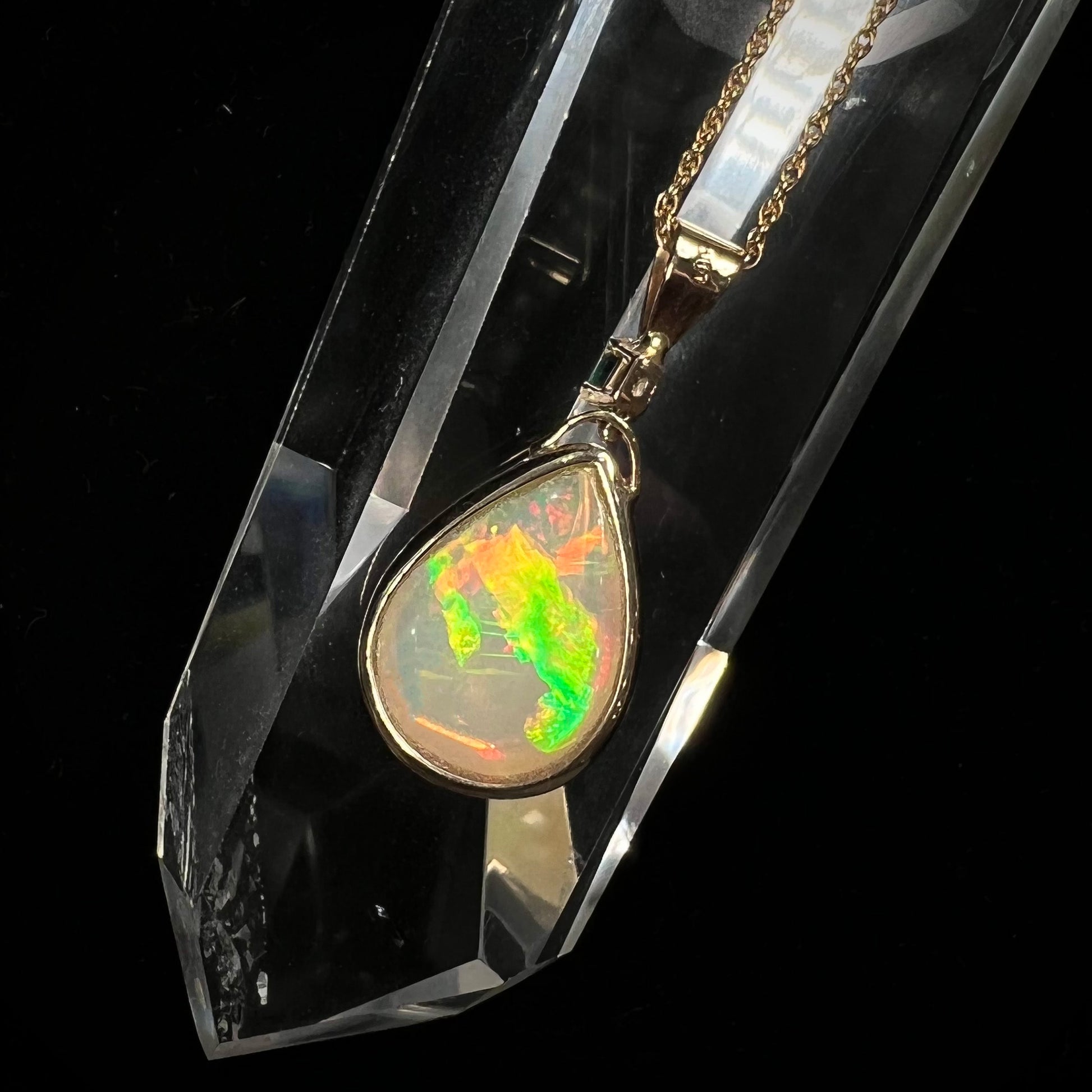 A yellow gold bezel set Ethiopian fire opal drop pendant.  There is an emerald set in the bail.
