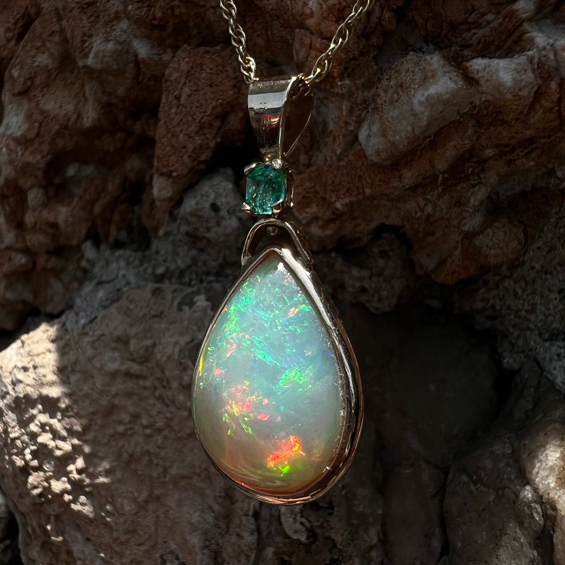 A yellow gold bezel set Ethiopian fire opal drop pendant.  There is an emerald set in the bail.
