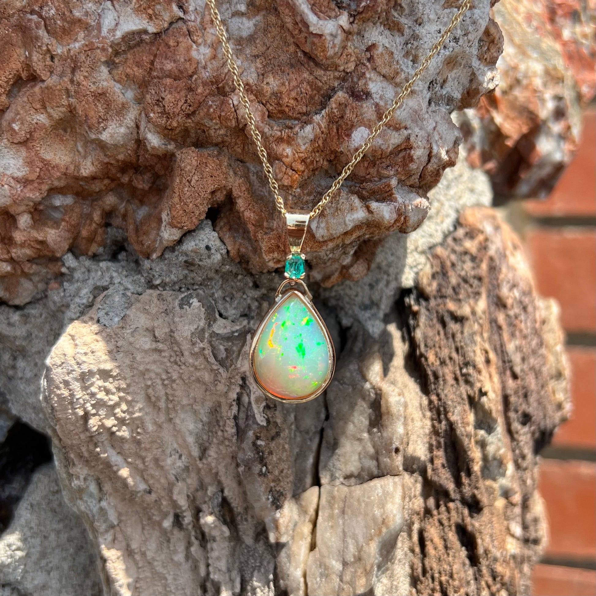 A yellow gold bezel set Ethiopian fire opal drop pendant.  There is an emerald set in the bail.