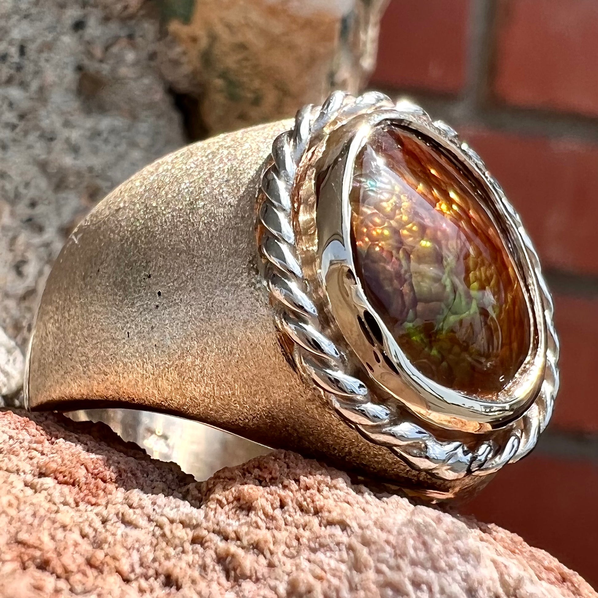 A men's matte finish textured gold fire agate ring.  There is a gold rope bezel around the oval cabochon cut stone.