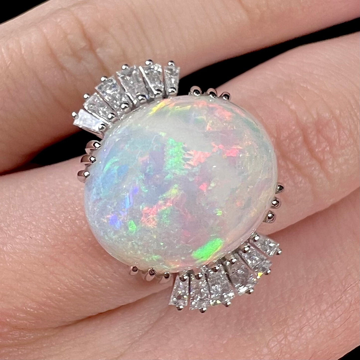 An oval cabochon cut opal mounted in a ladies' white gold ring between tapered baguette cut diamonds.