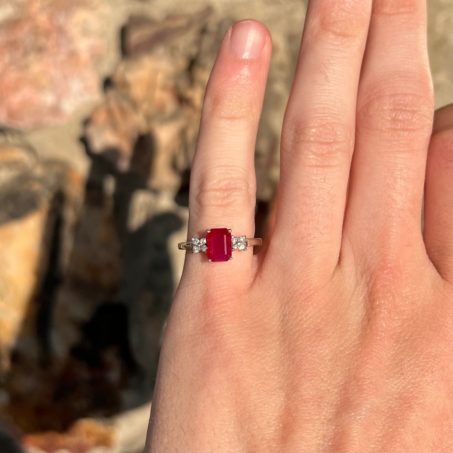 A white gold ring with an emerald cut red synthetic ruby set between four round cut diamonds on each side.