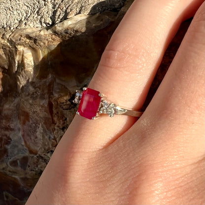 A white gold ring with an emerald cut red synthetic ruby set between four round cut diamonds on each side.