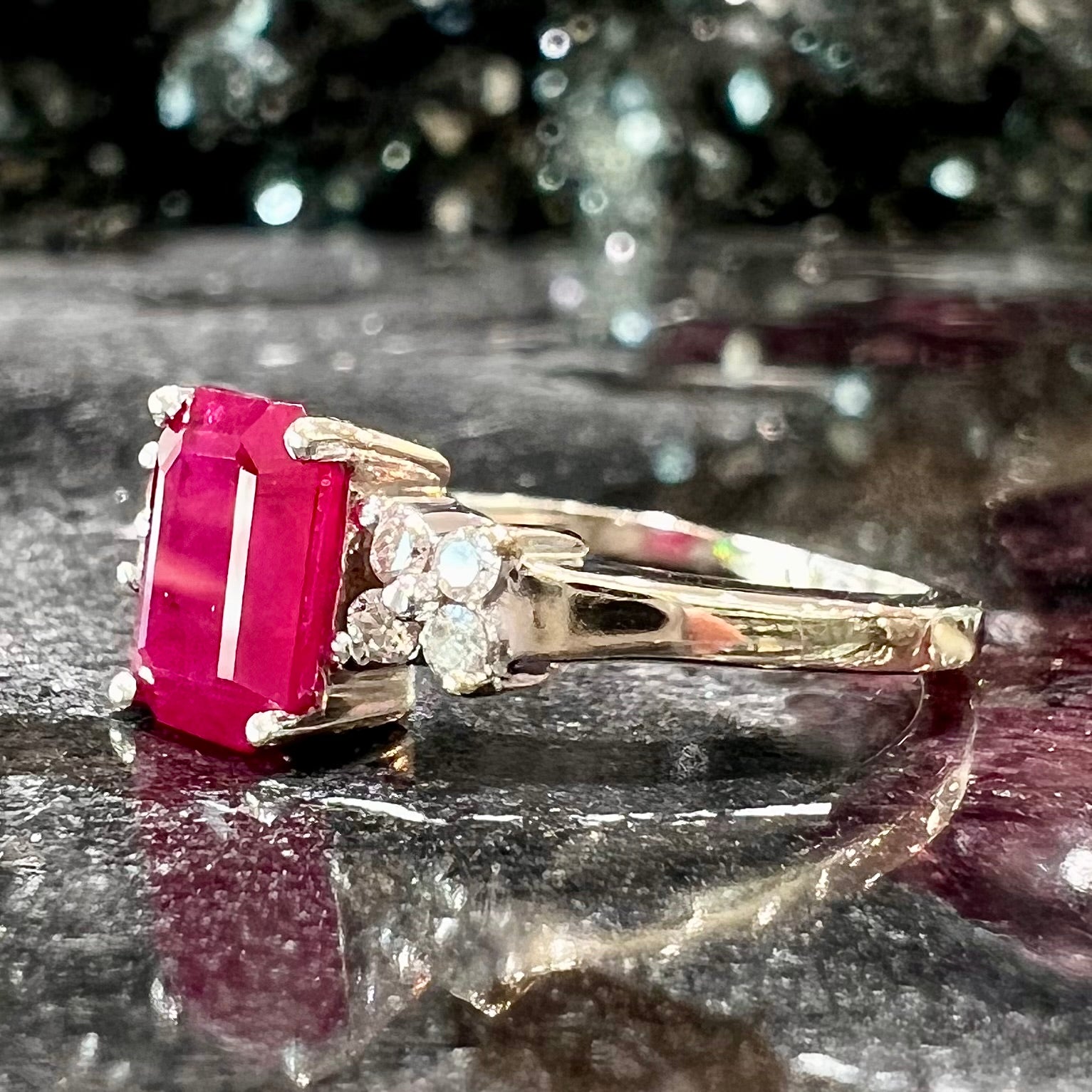 A white gold ring with an emerald cut red synthetic ruby set between four round cut diamonds on each side.