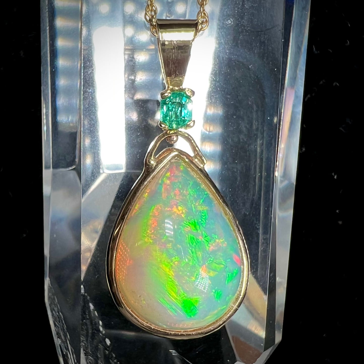 A yellow gold bezel set Ethiopian fire opal drop pendant.  There is an emerald set in the bail.