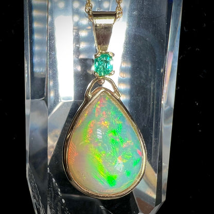 A yellow gold bezel set Ethiopian fire opal drop pendant.  There is an emerald set in the bail.