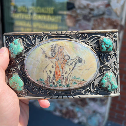 A men's Navajo-made white brass belt buckle set with turquoise nuggets and a mother of pearl stone etched with a Native American riding a horse.