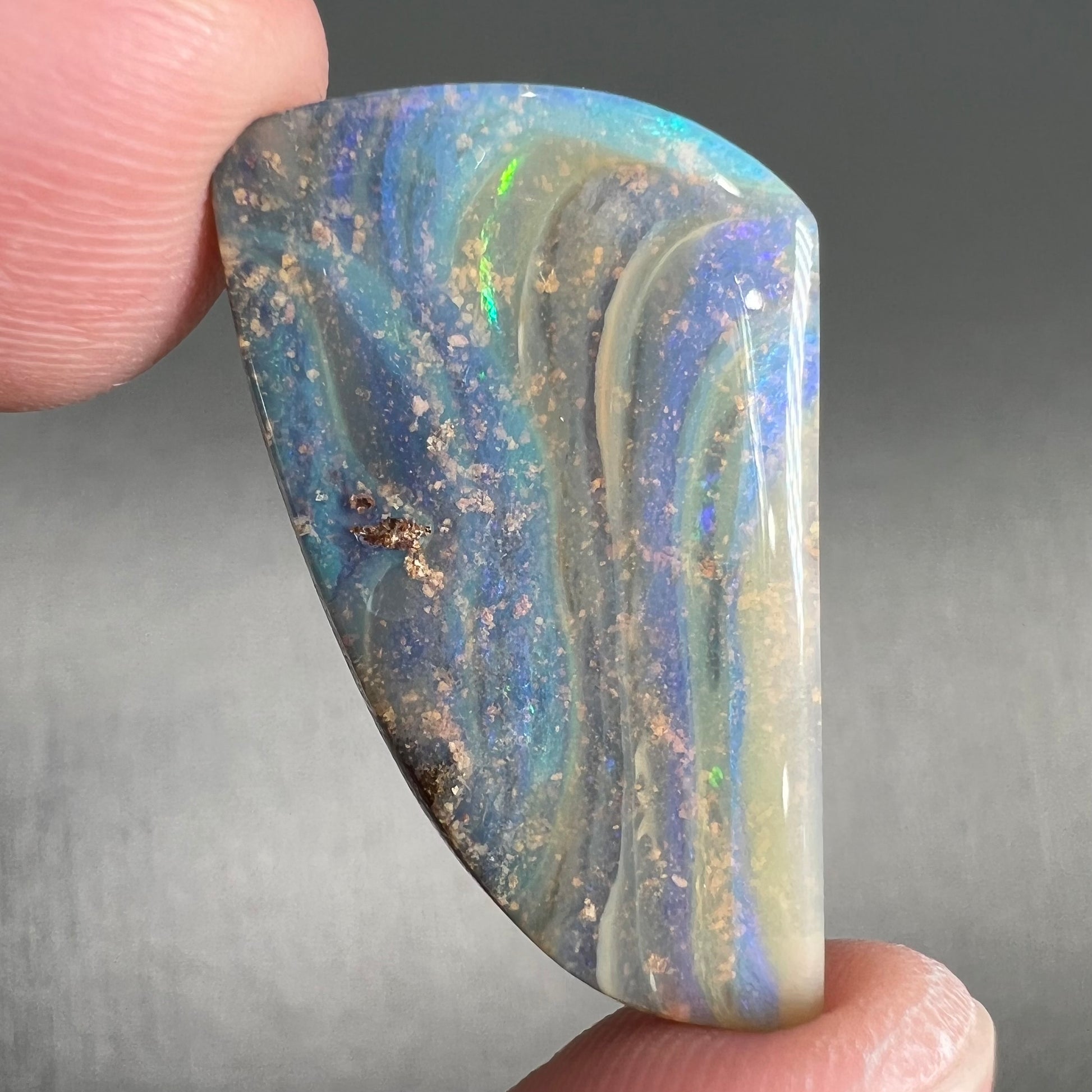 A polished, freeform shaped boulder opal stone from Queensland, Australia.  Predominant colors are green, blue, and violet.