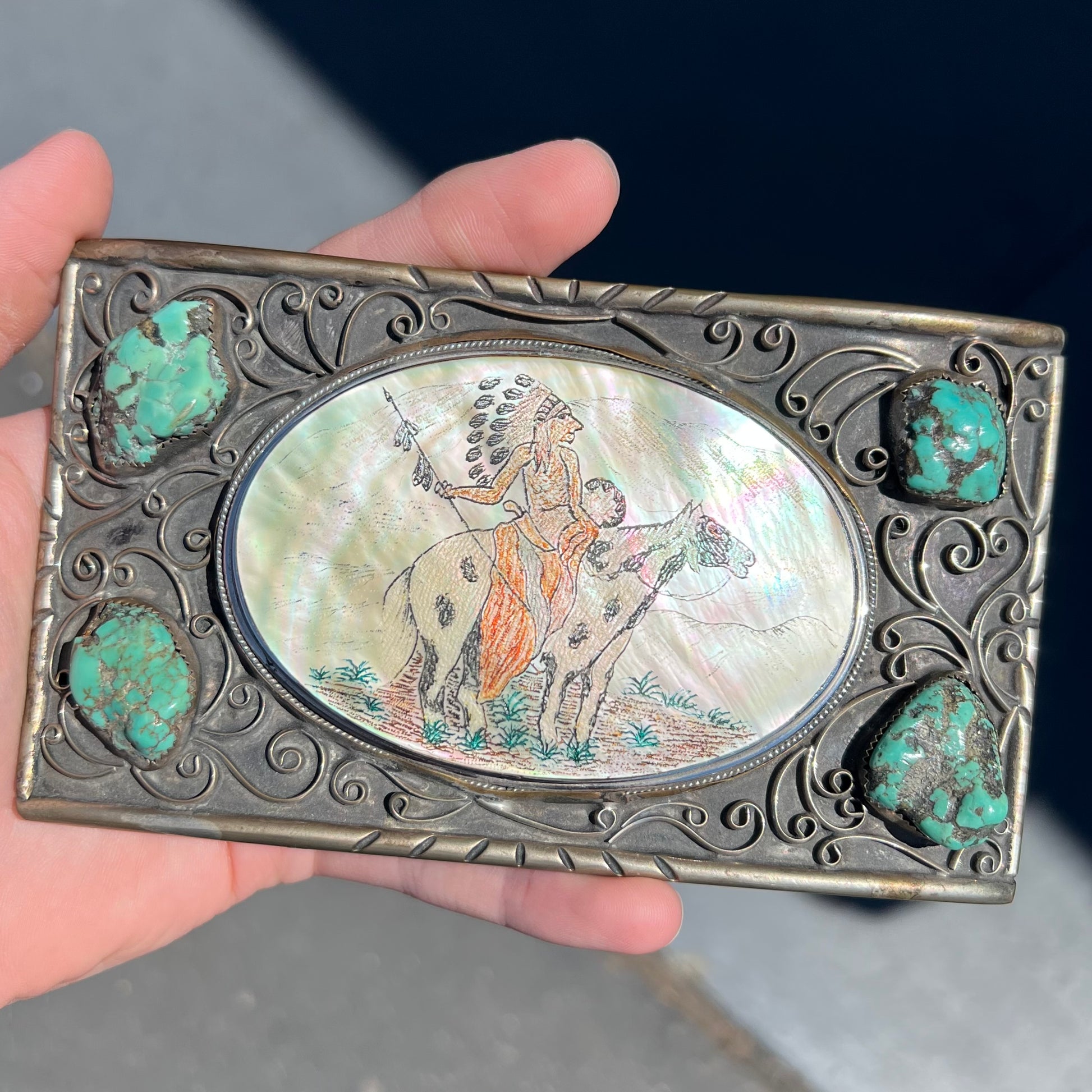 A men's Navajo-made white brass belt buckle set with turquoise nuggets and a mother of pearl stone etched with a Native American riding a horse.