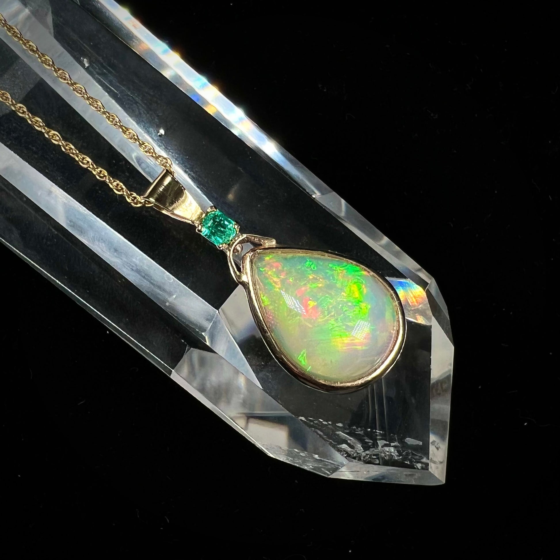A yellow gold bezel set Ethiopian fire opal drop pendant.  There is an emerald set in the bail.