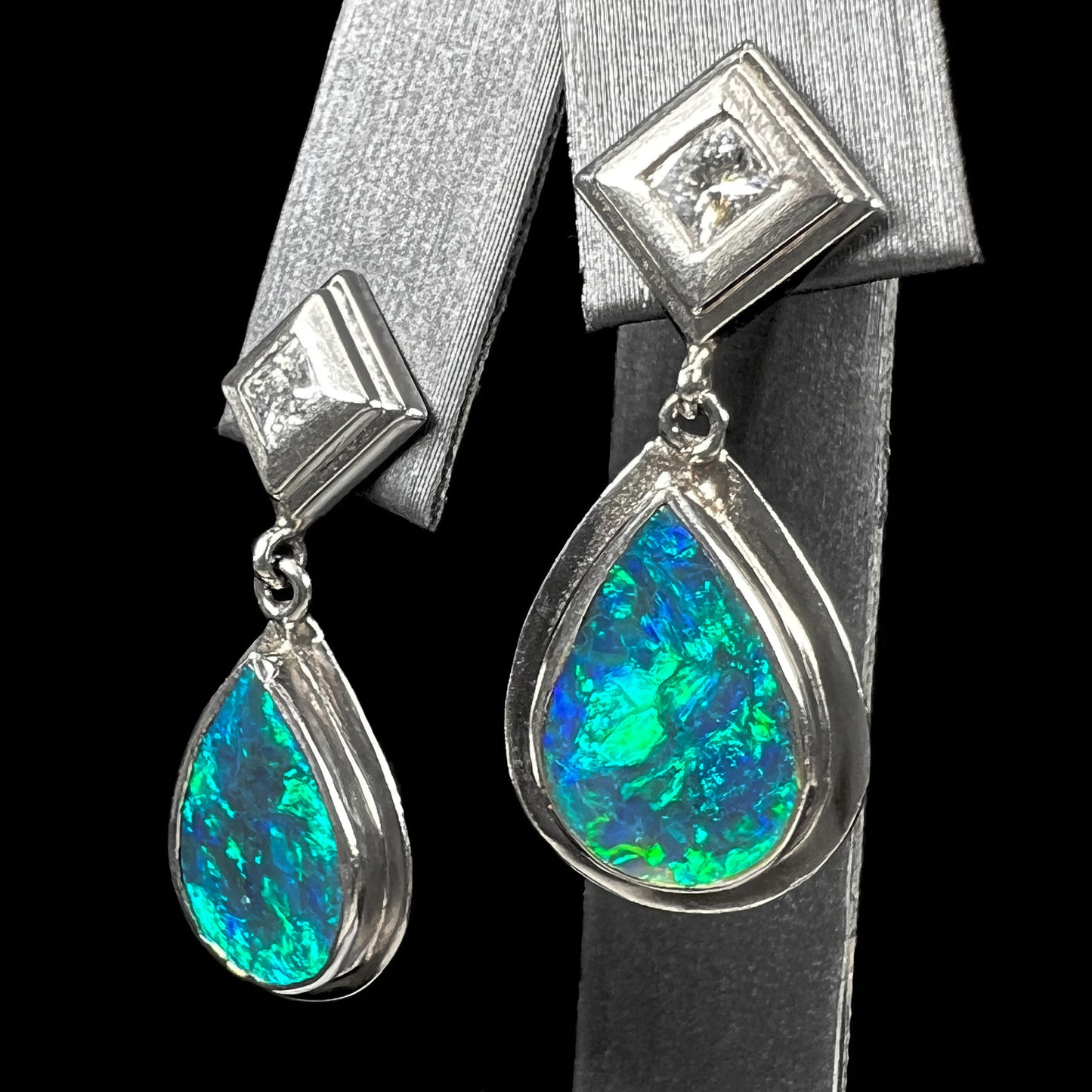 Black crystal opal dangle earrings with blue body color and green play of color set with princess cut diamonds in platinum.