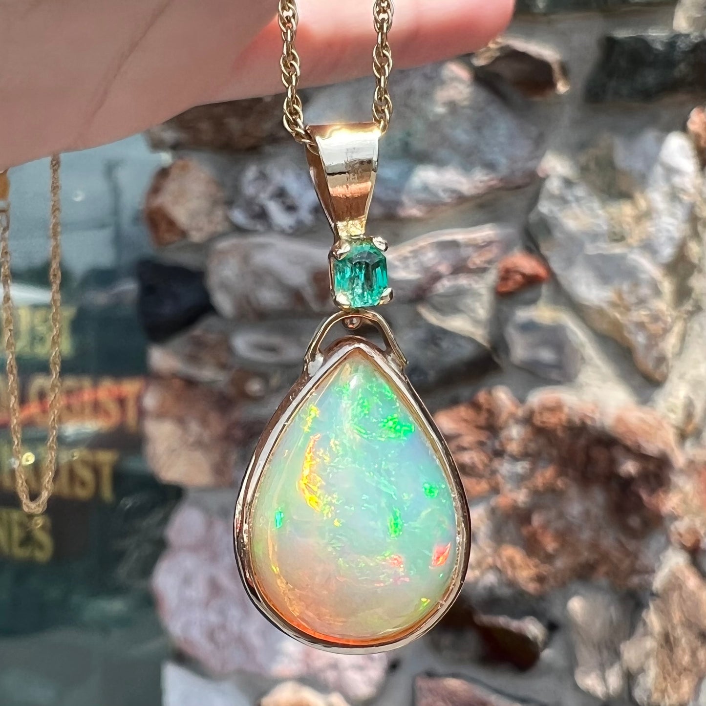 A yellow gold bezel set Mexican fire opal drop pendant.  There is an emerald set in the bail.