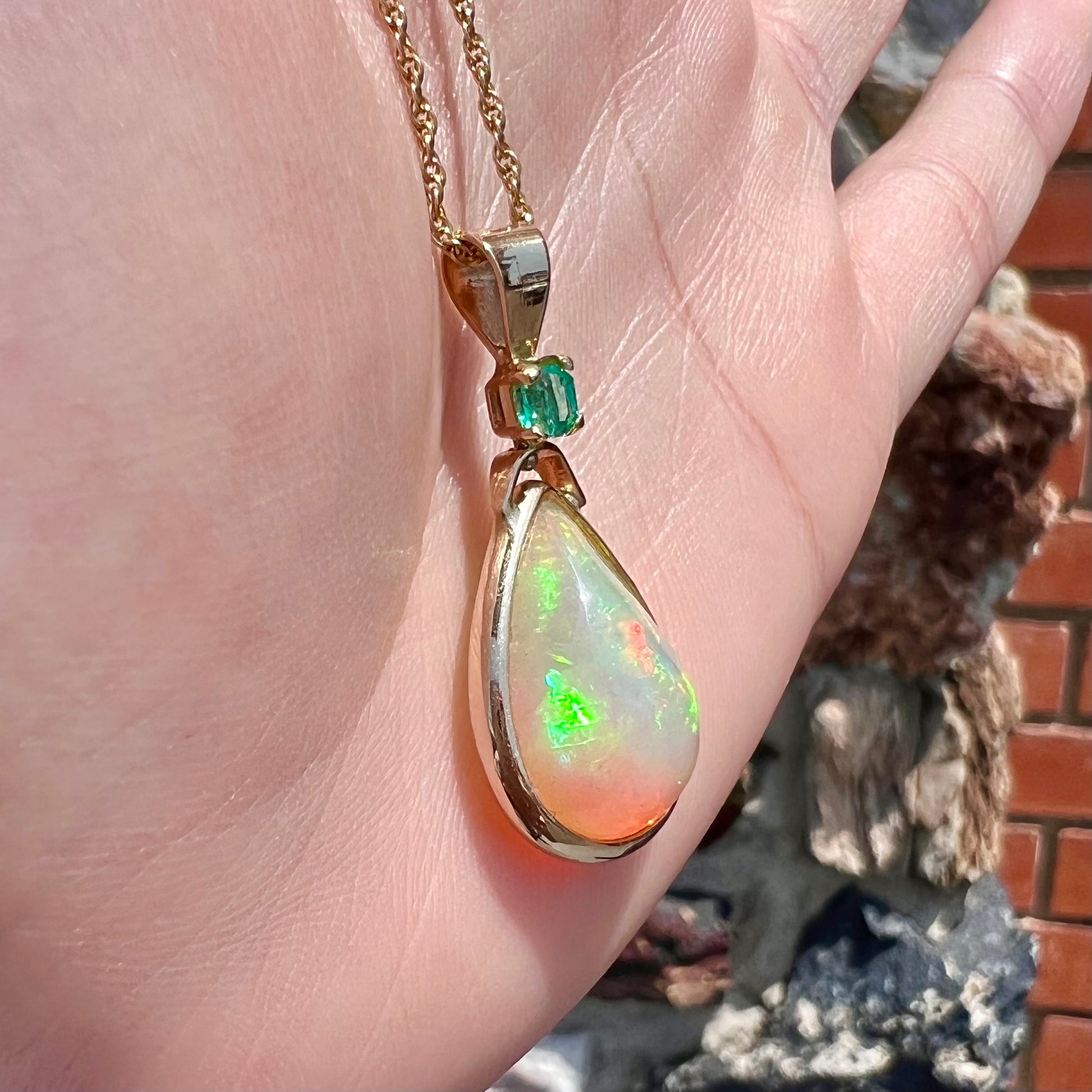 A yellow gold bezel set Ethiopian fire opal drop pendant.  There is an emerald set in the bail.