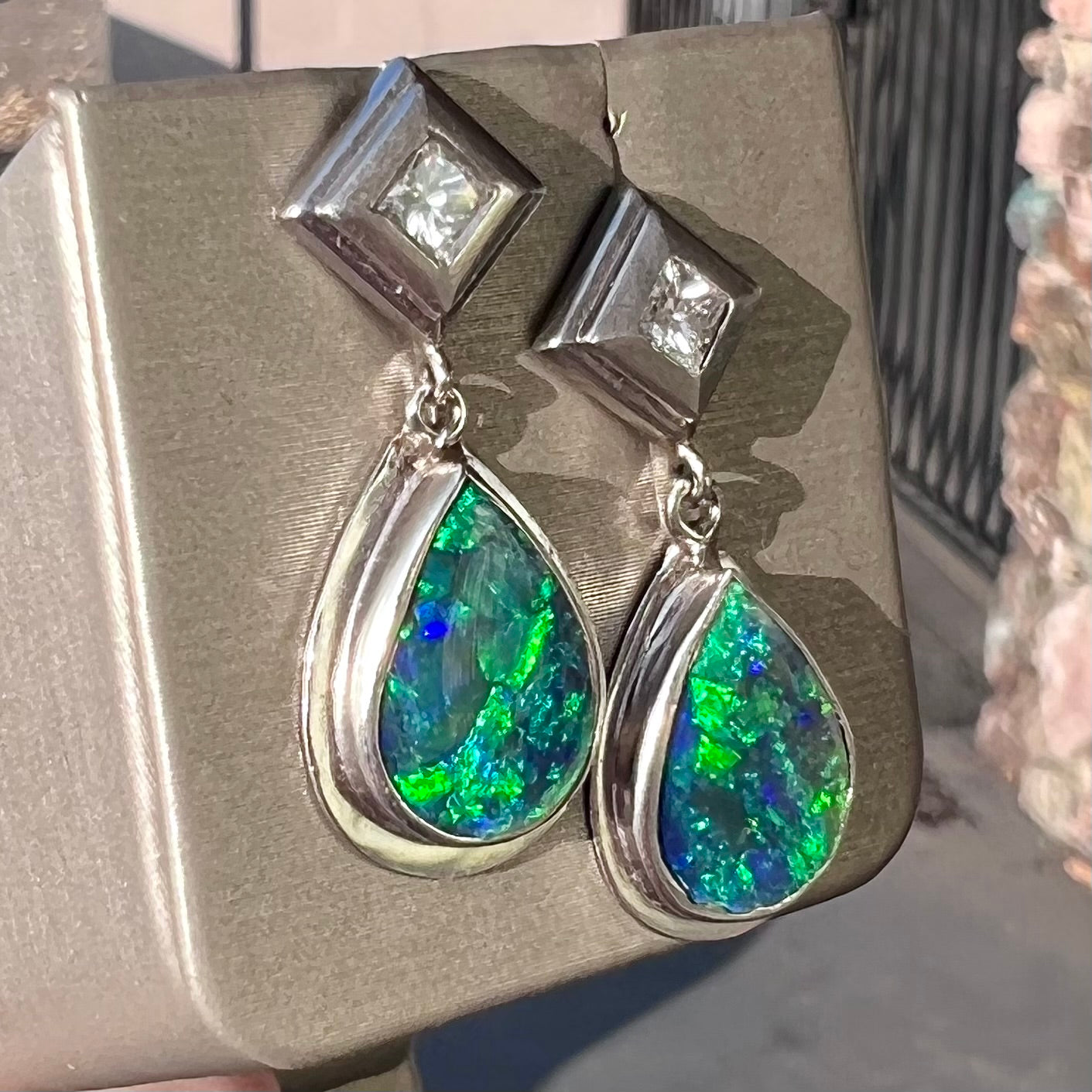 Black crystal opal dangle earrings with blue body color and green play of color set with princess cut diamonds in platinum.