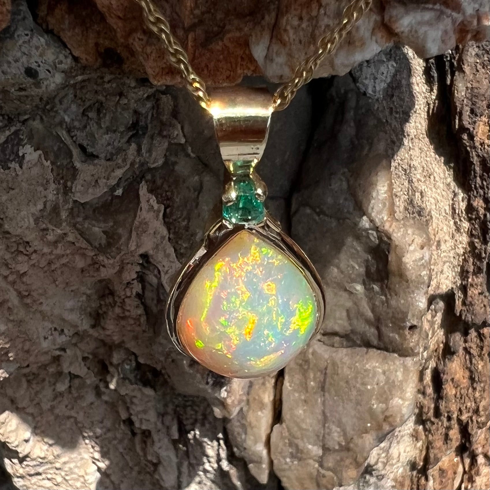 A yellow gold bezel set Ethiopian fire opal drop pendant.  There is an emerald set in the bail.