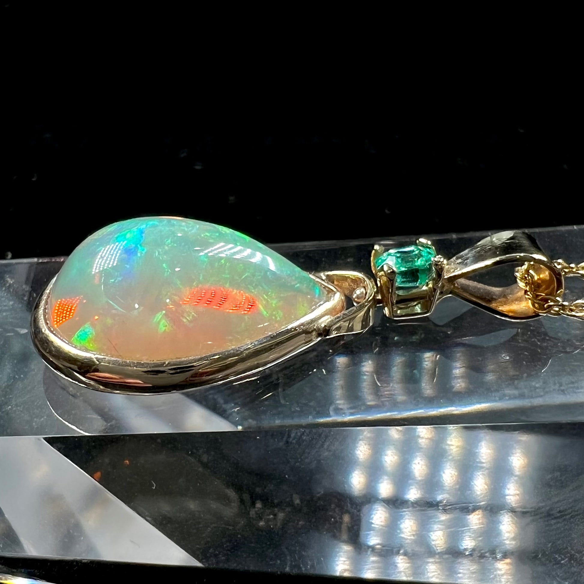 A yellow gold bezel set Ethiopian fire opal drop pendant.  There is an emerald set in the bail.