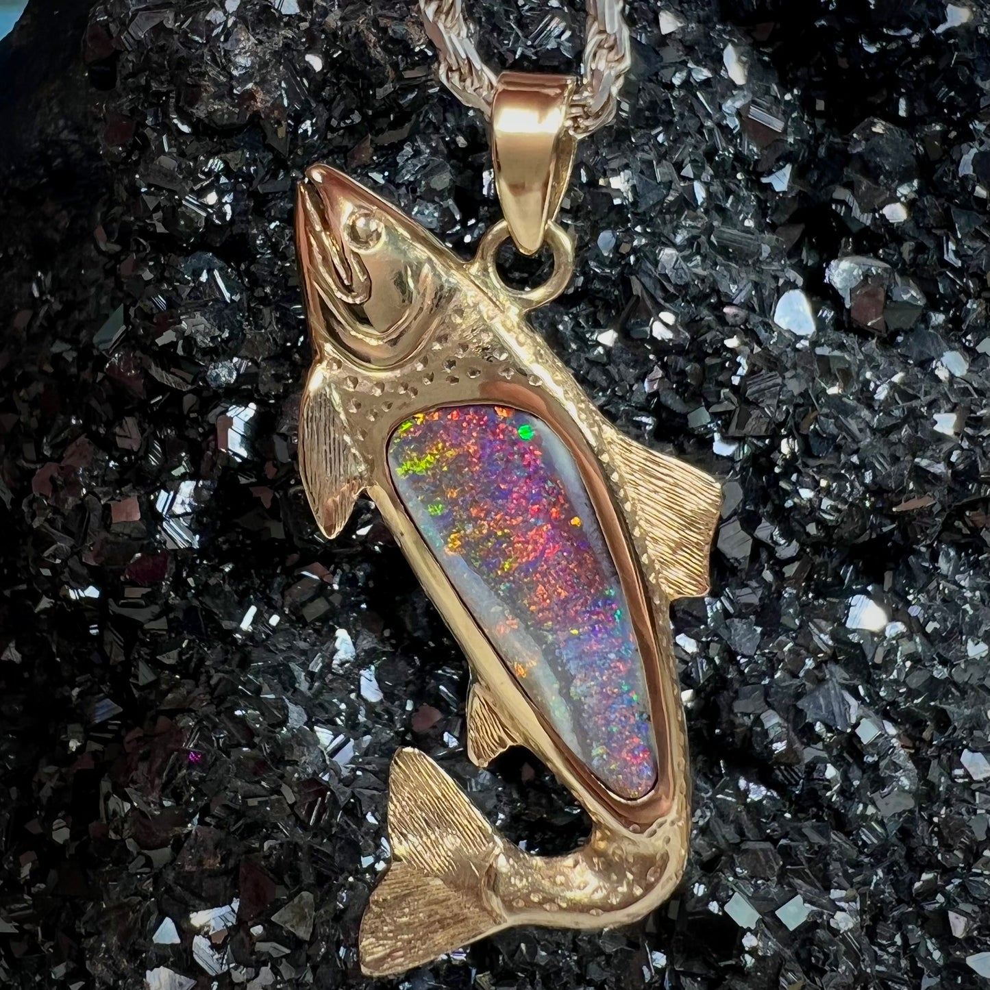 A yellow gold fish pendant detailed to look like a rainbow trout.  The body of the fish is set with a color changing black opal stone.