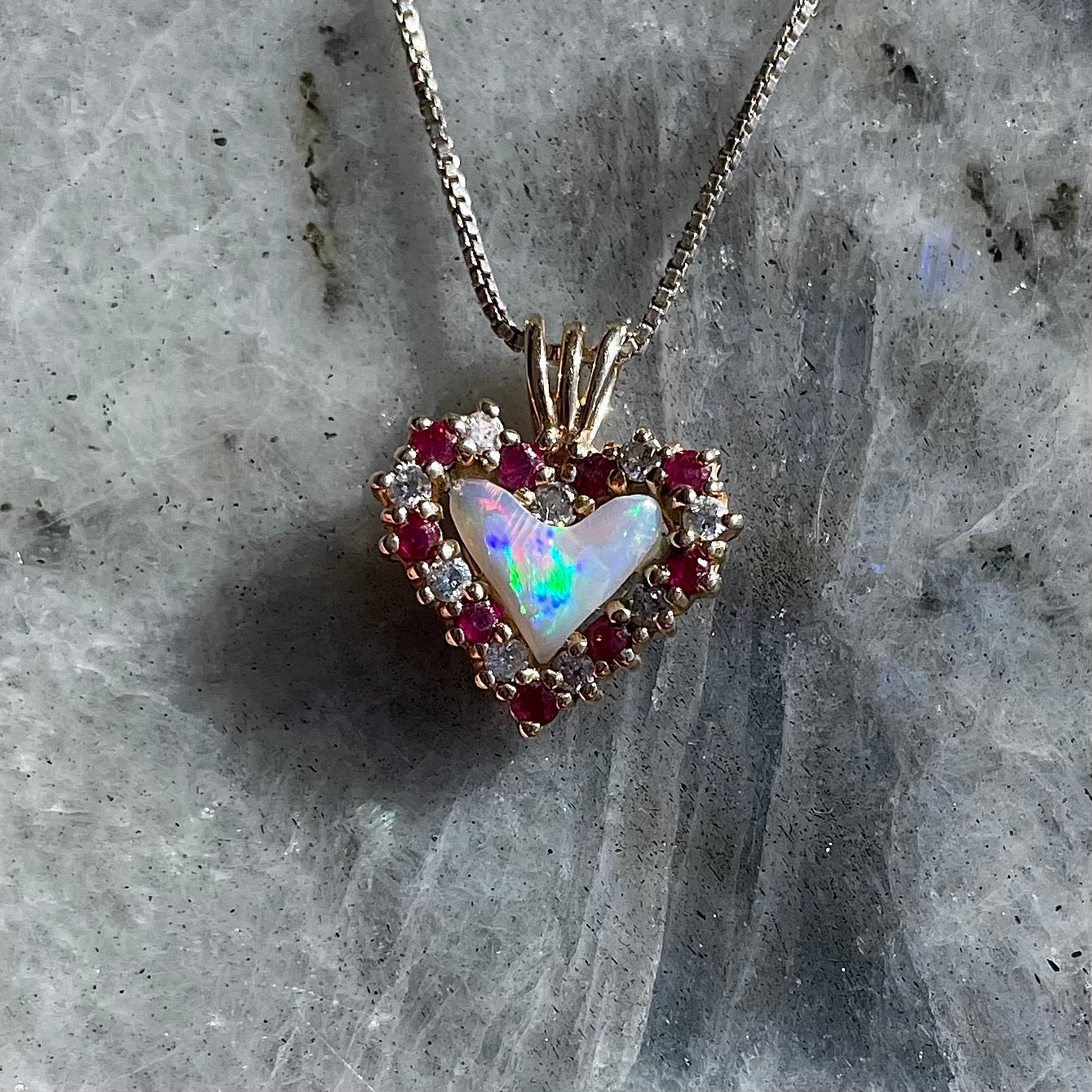 A yellow gold pendant set with a heart cut white opal in a halo of alternating round rubies and diamonds.
