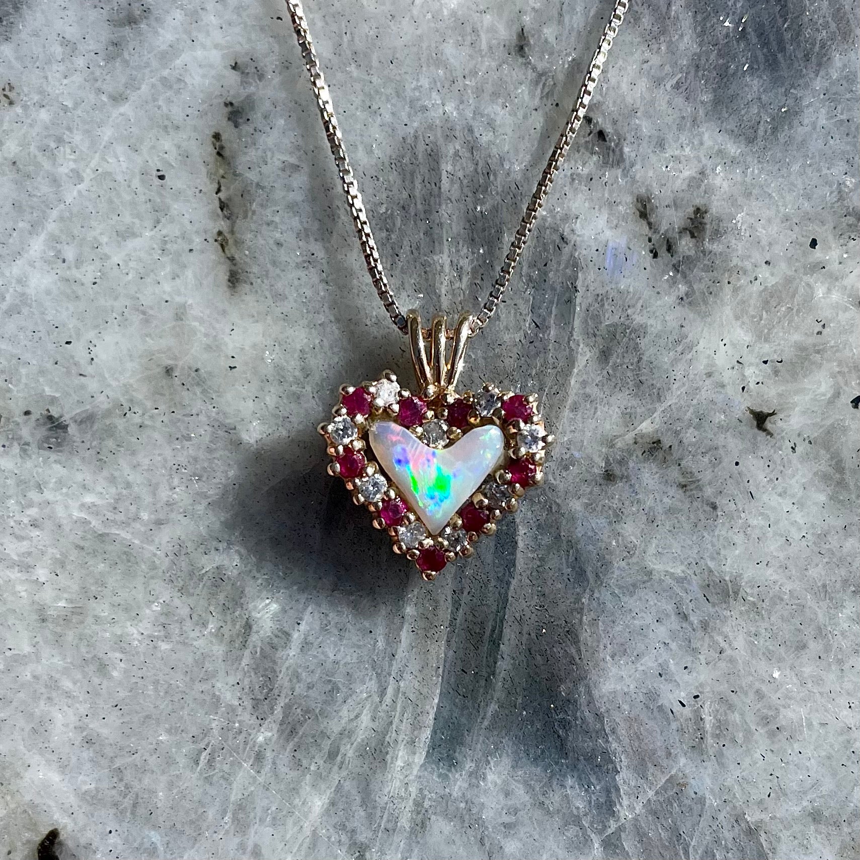 A yellow gold pendant set with a heart cut white opal in a halo of alternating round rubies and diamonds.