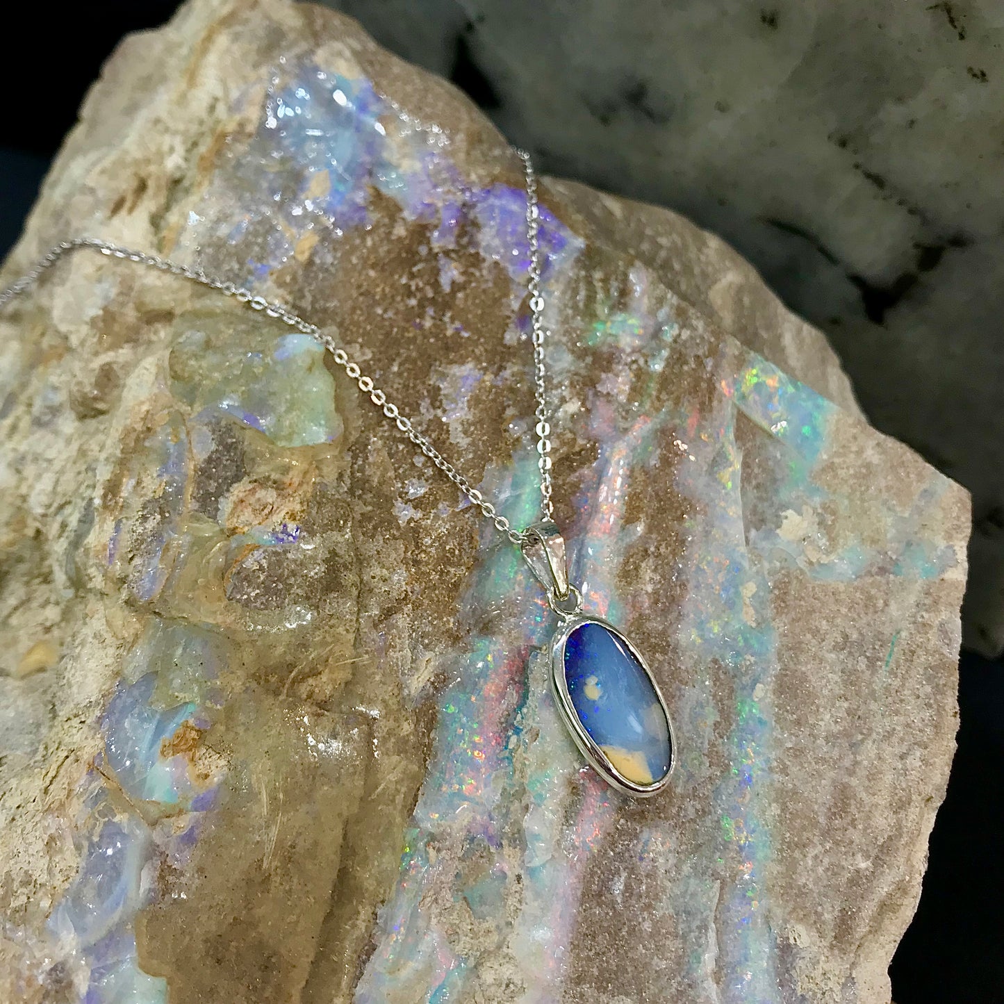 A photo of a Queensland Australian blue boulder opal with tan matrix set into a sterling silver bezel pendant.  The opal resembles a beach with tan sand matrix and a blue ocean of color.  The pendant is on a sterling silver cable chain and against a natural rough white crystal opal specimen backdrop.