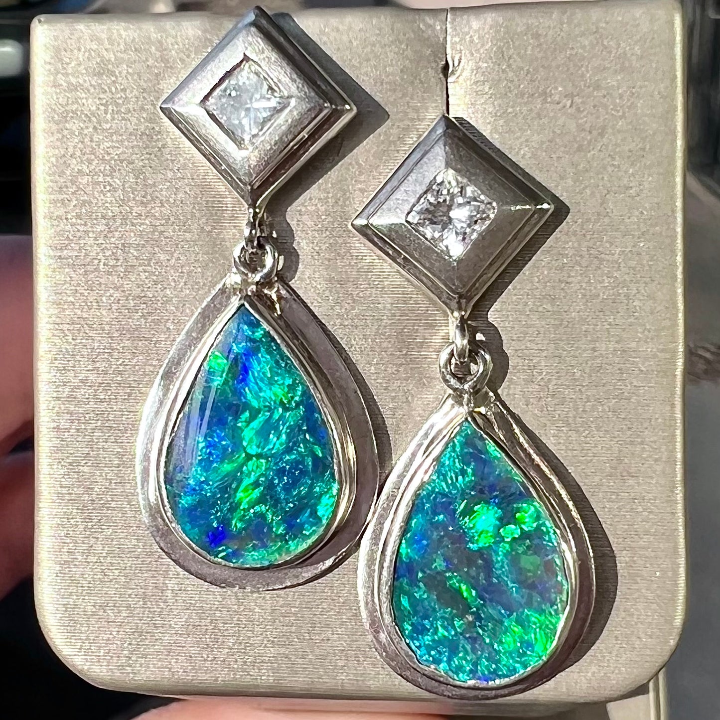 Black crystal opal dangle earrings with blue body color and green play of color set with princess cut diamonds in platinum.