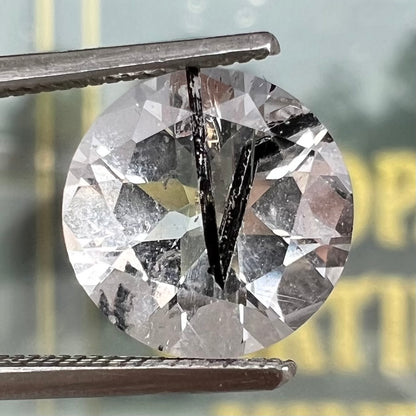 A faceted round brilliant cut clear quartz stone with black tourmaline inclusions.  Inclusions appear like a clock's hands.