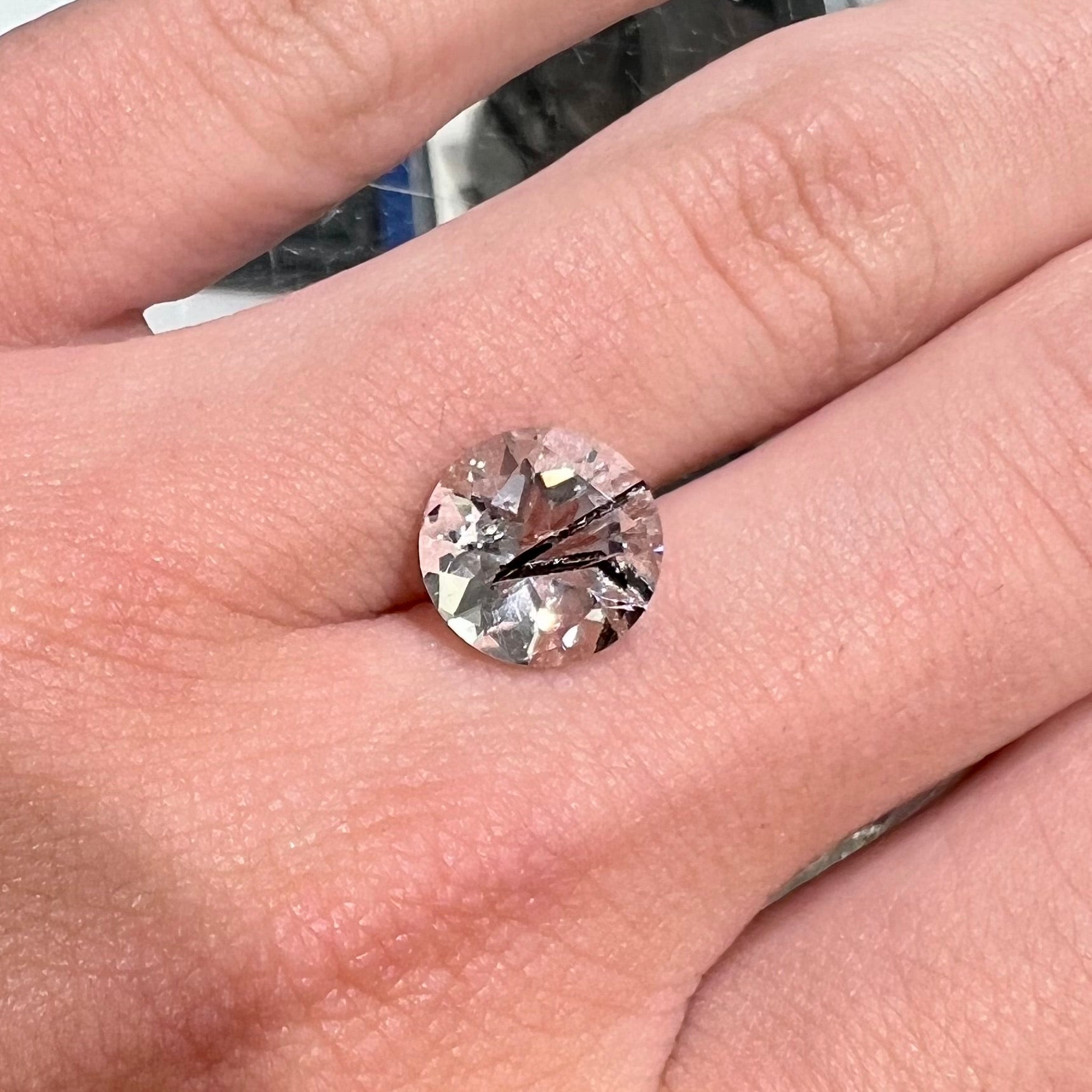 A faceted round brilliant cut clear quartz stone with black tourmaline inclusions.  Inclusions appear like a clock's hands.