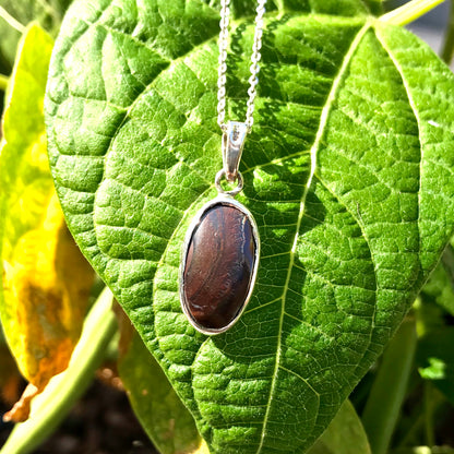 This photo shows the backside of an Australian boulder opal pendant set in sterling silver.  The backside of the opal shows its banded brown ironstone matrix.  The piece is on a sterling silver cable chain and against a green leaf backdrop.