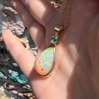 A yellow gold bezel set Ethiopian fire opal drop pendant.  There is an emerald set in the bail.