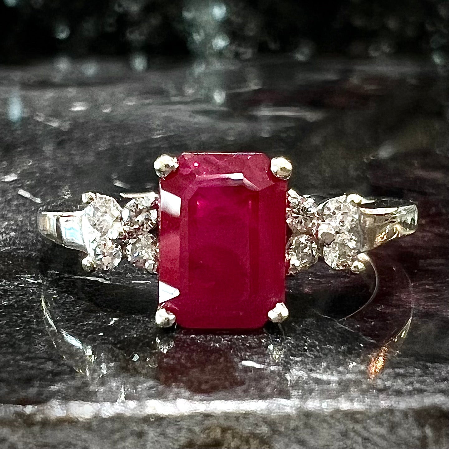 A white gold ring with an emerald cut red synthetic ruby set between four round cut diamonds on each side.