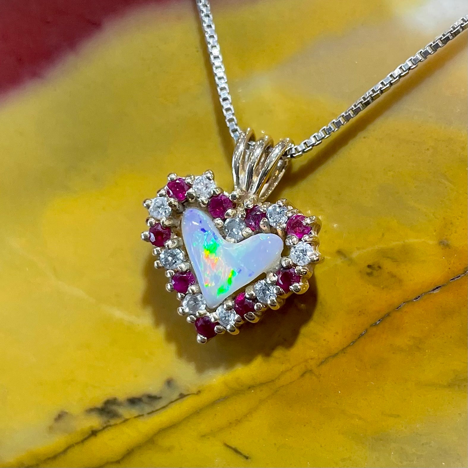 A yellow gold pendant set with a heart cut white opal in a halo of alternating round rubies and diamonds.