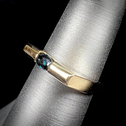 A ladies' yellow gold solitire ring horizontally set with an oval cut dark teal blue sapphire stone.