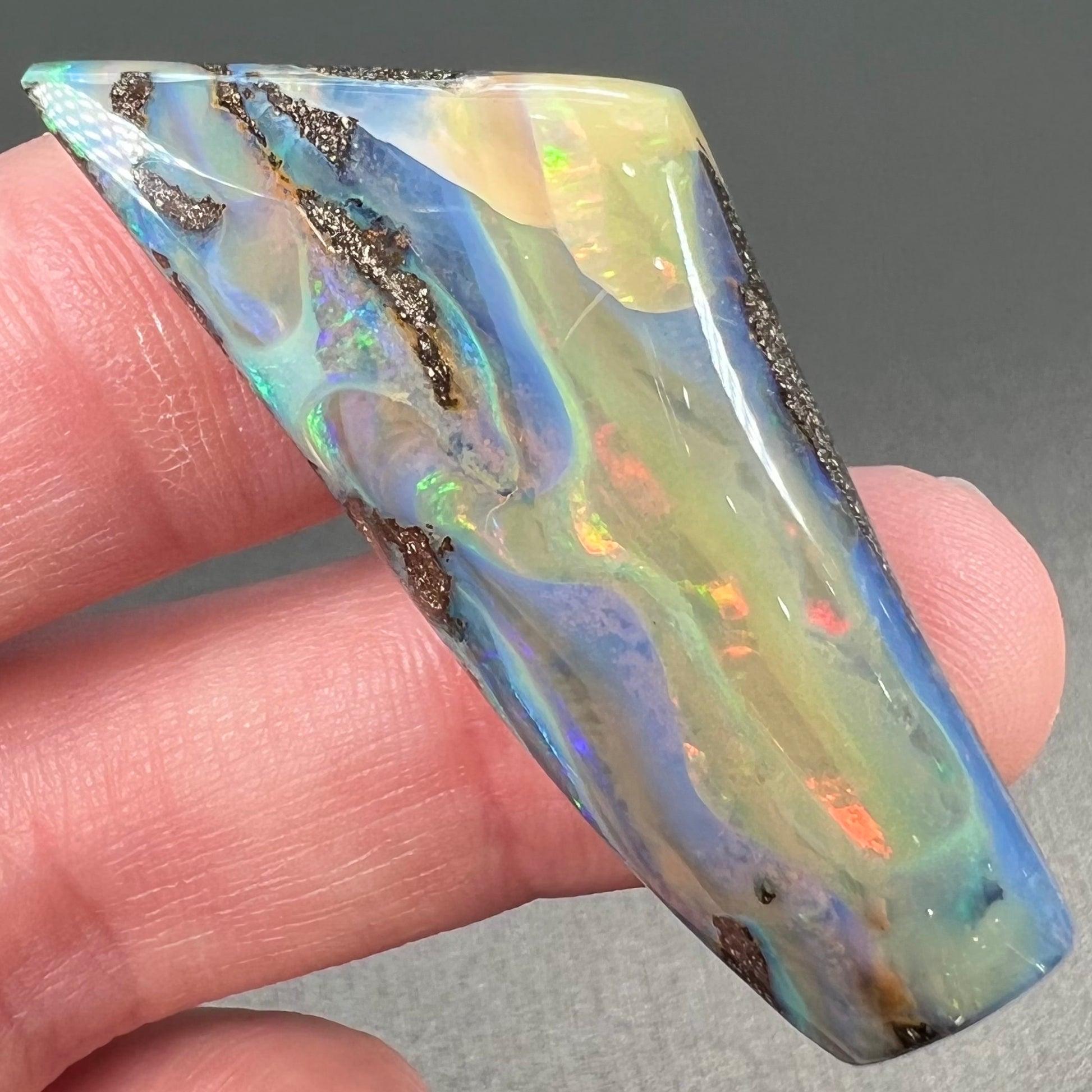 A freeform shaped, polished boulder opal stone from Queensland, Australia.  The stone has red, orange, green, and blue fire.