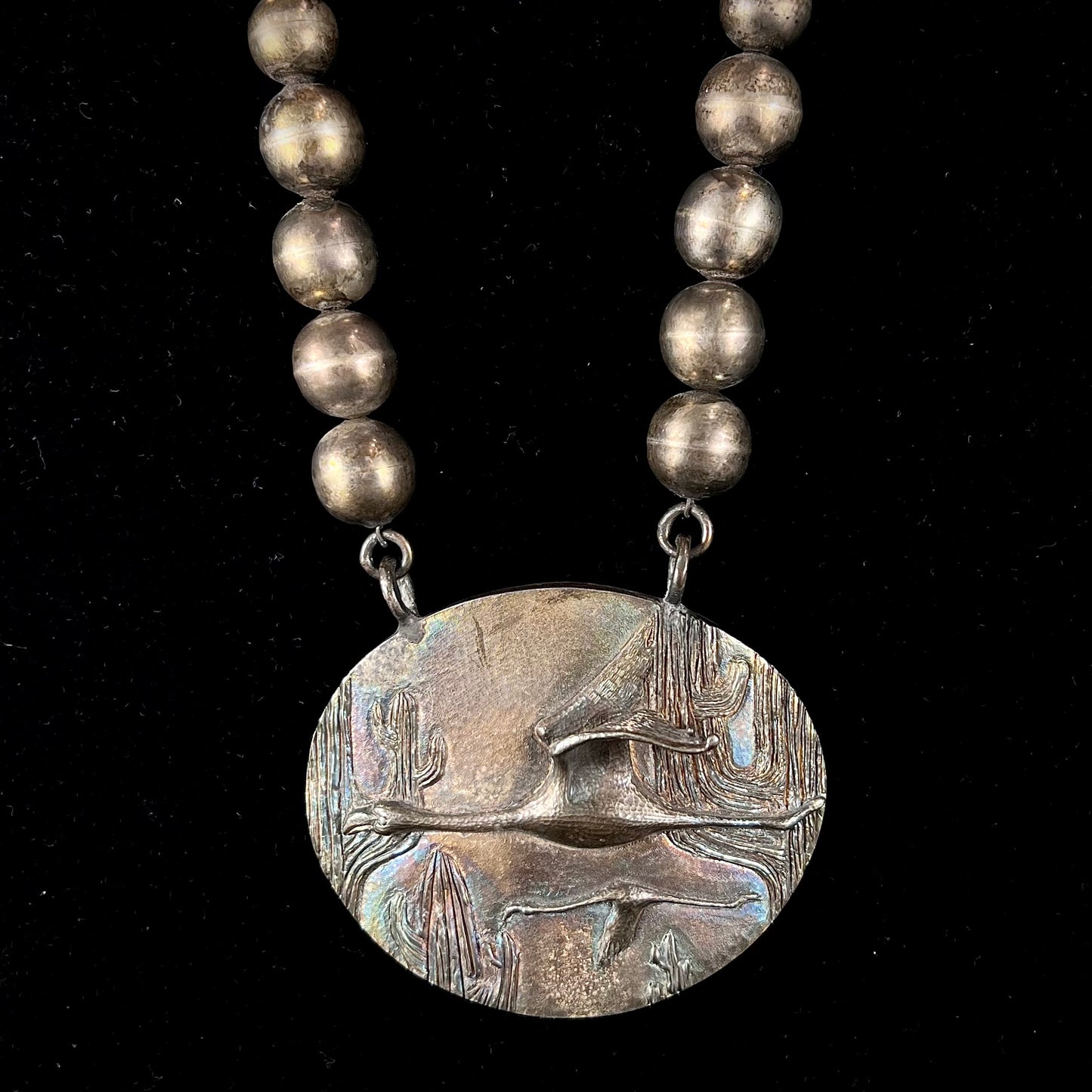 A handmade sterling silver bead necklace with a rainbow patinaed medallion that features 3D cast flamingos flying through a cactus desert.