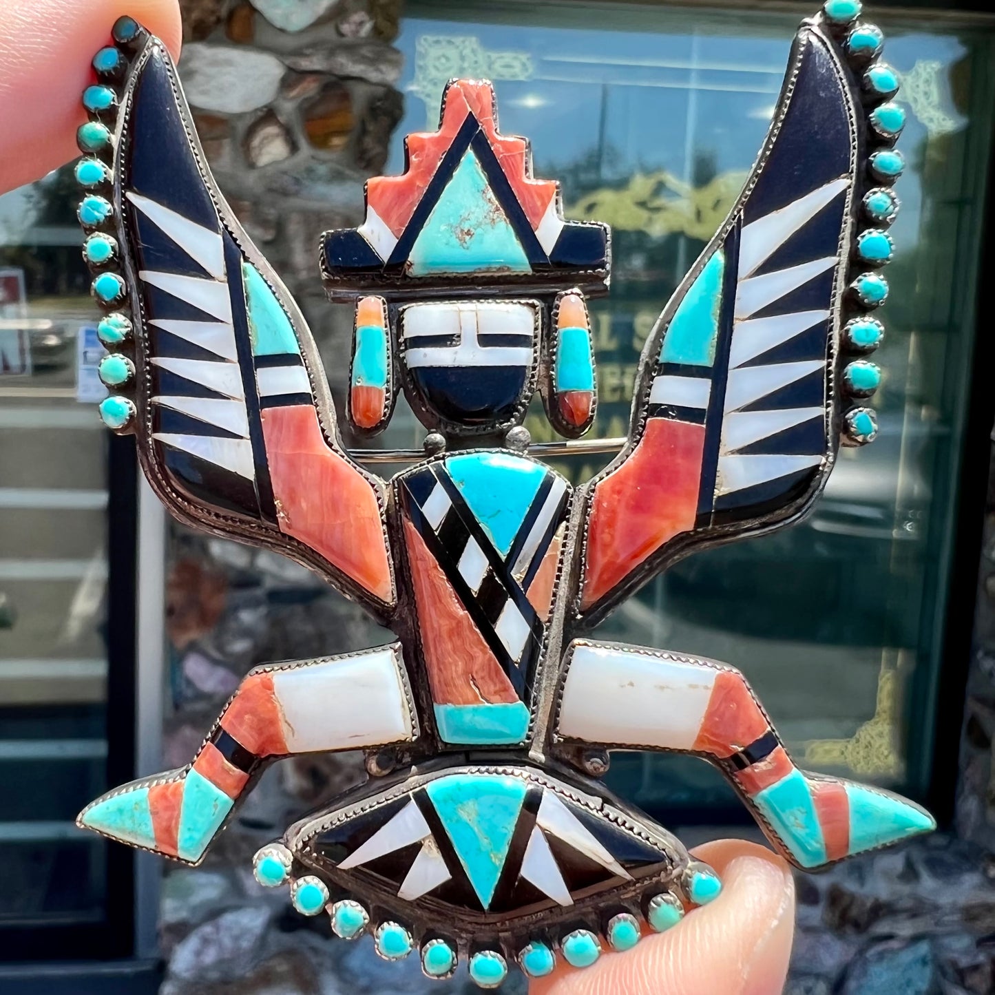 A handmade, silver Zuni dancing eagle pin.  The piece is inlaid with turquoise, coral, mother of pearl, and onyx stones.