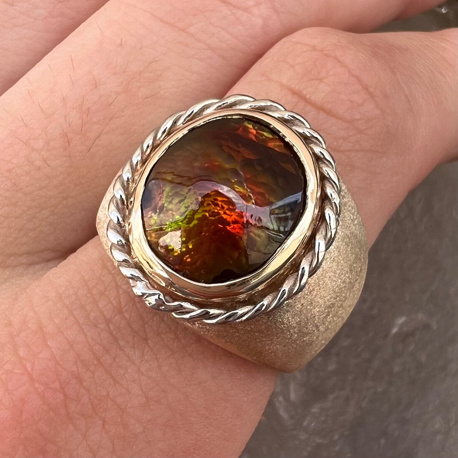 A men's matte finish textured gold fire agate ring.  There is a gold rope bezel around the oval cabochon cut stone.