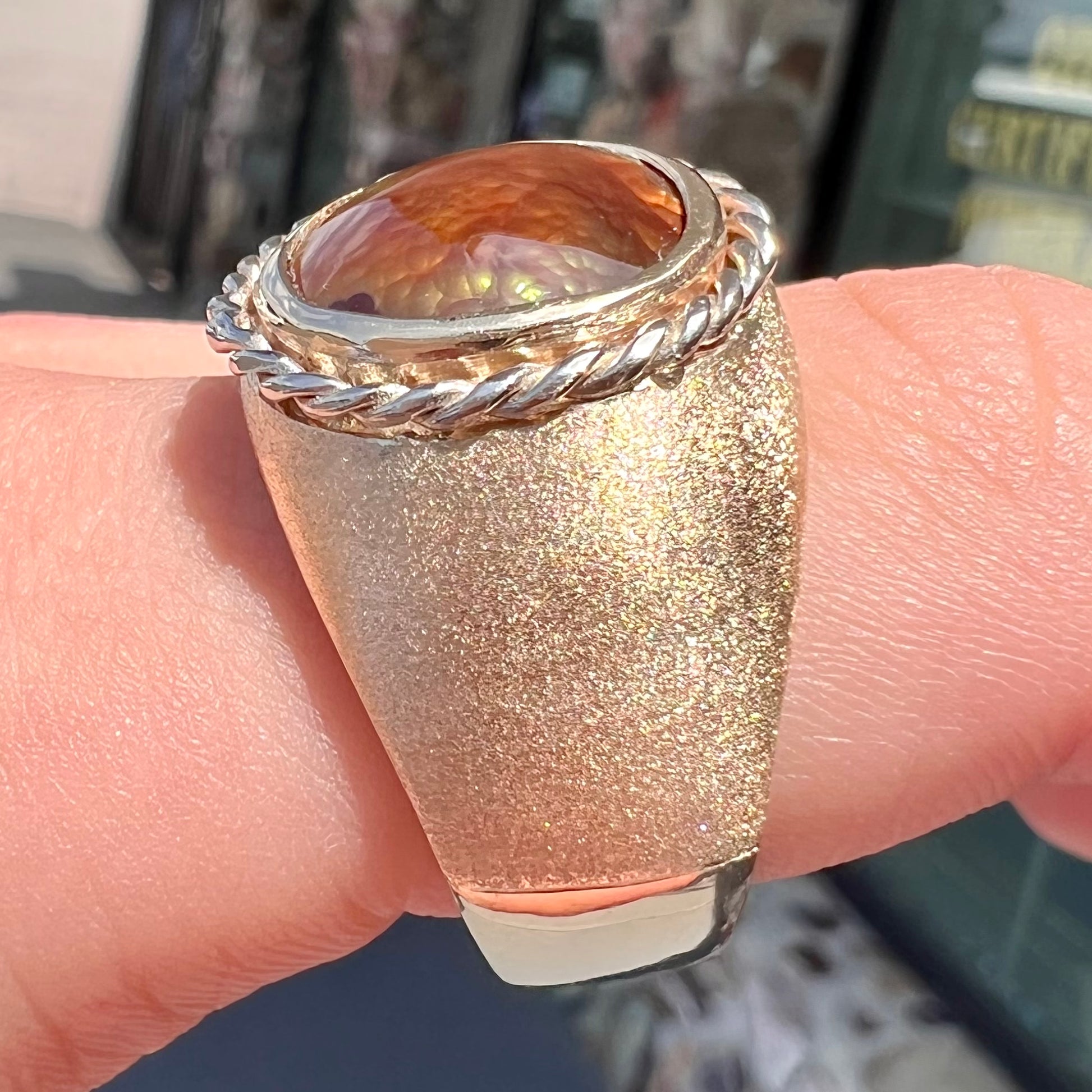 A men's matte finish textured gold fire agate ring.  There is a gold rope bezel around the oval cabochon cut stone.