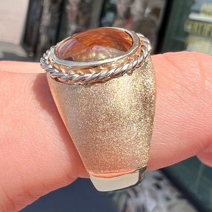 A men's matte finish textured gold fire agate ring.  There is a gold rope bezel around the oval cabochon cut stone.
