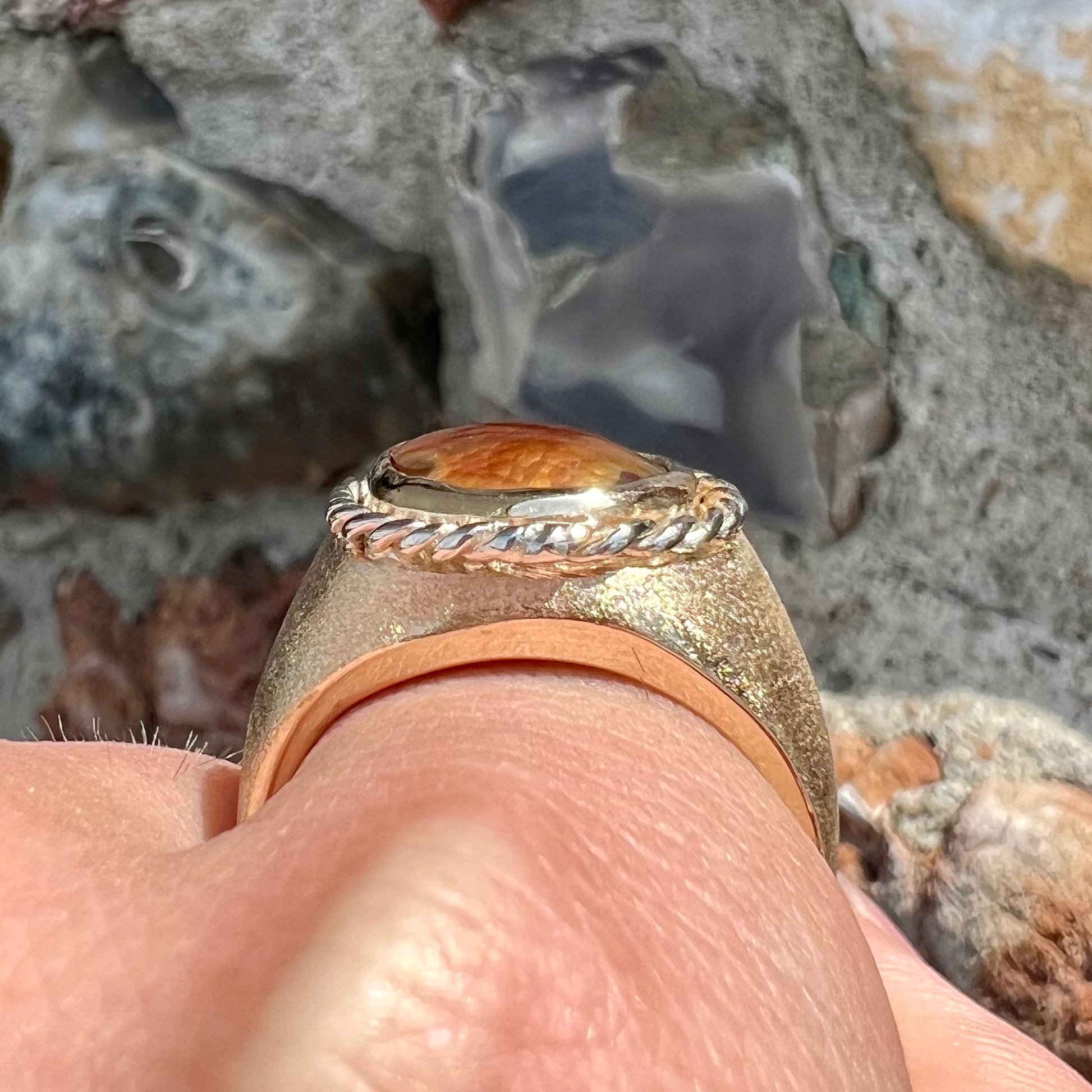 A men's matte finish textured gold fire agate ring.  There is a gold rope bezel around the oval cabochon cut stone.