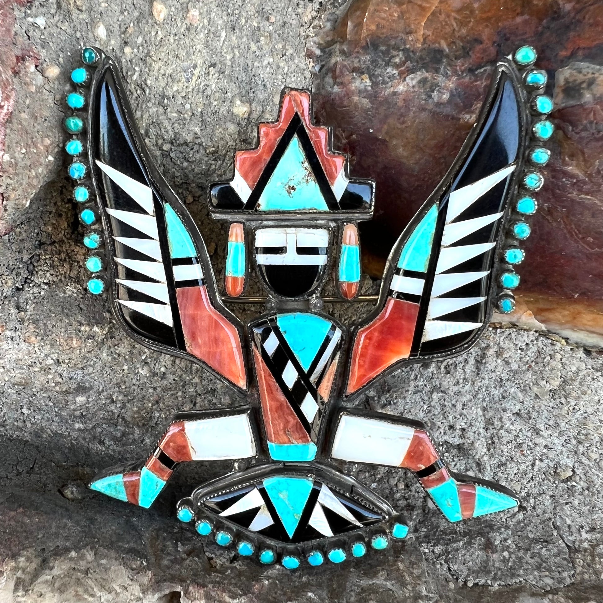 A handmade, silver Zuni dancing eagle pin.  The piece is inlaid with turquoise, coral, mother of pearl, and onyx stones.