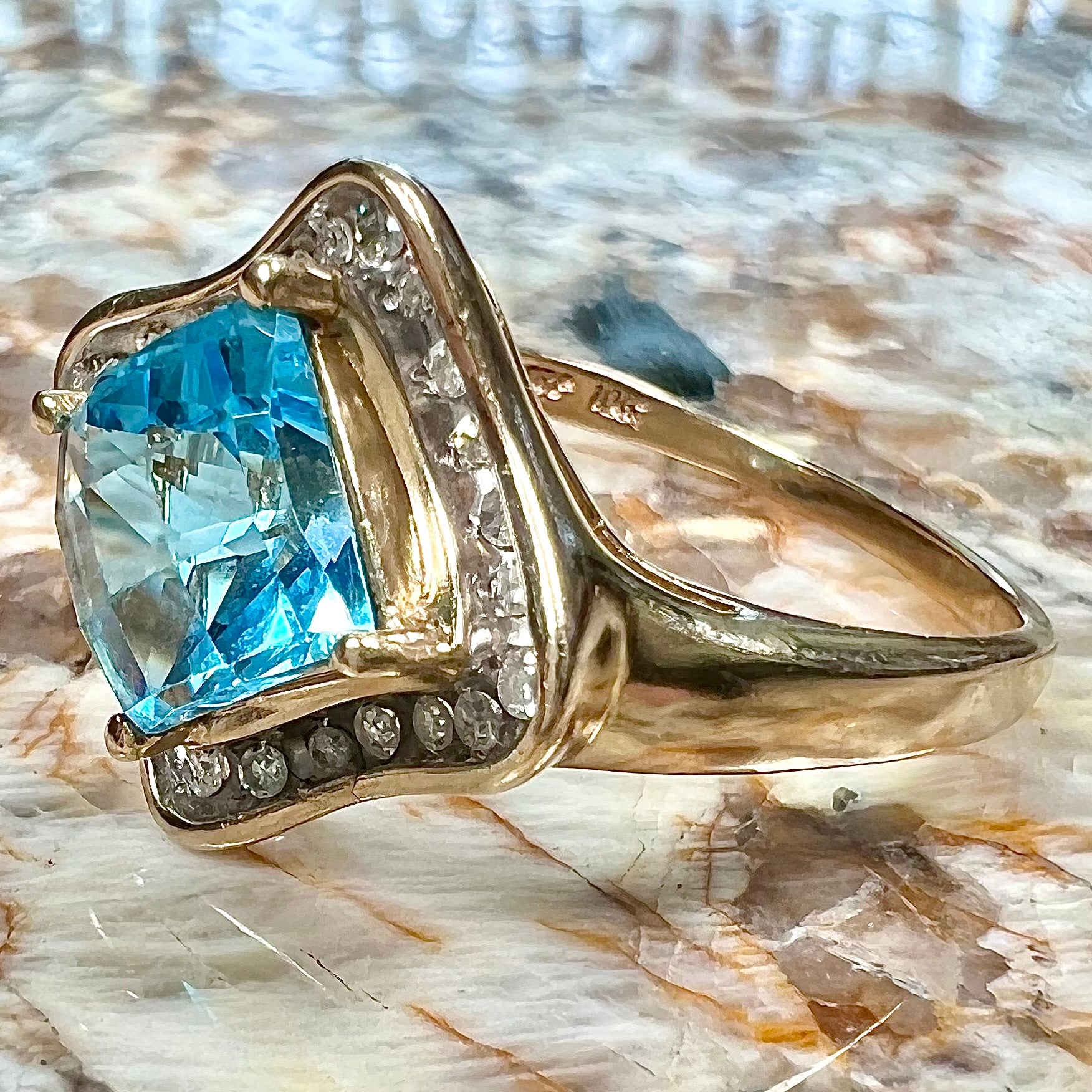 A checkerboard princess cut Swiss blue topaz ring.  The stone is set in a yellow gold, diamond halo mounting.