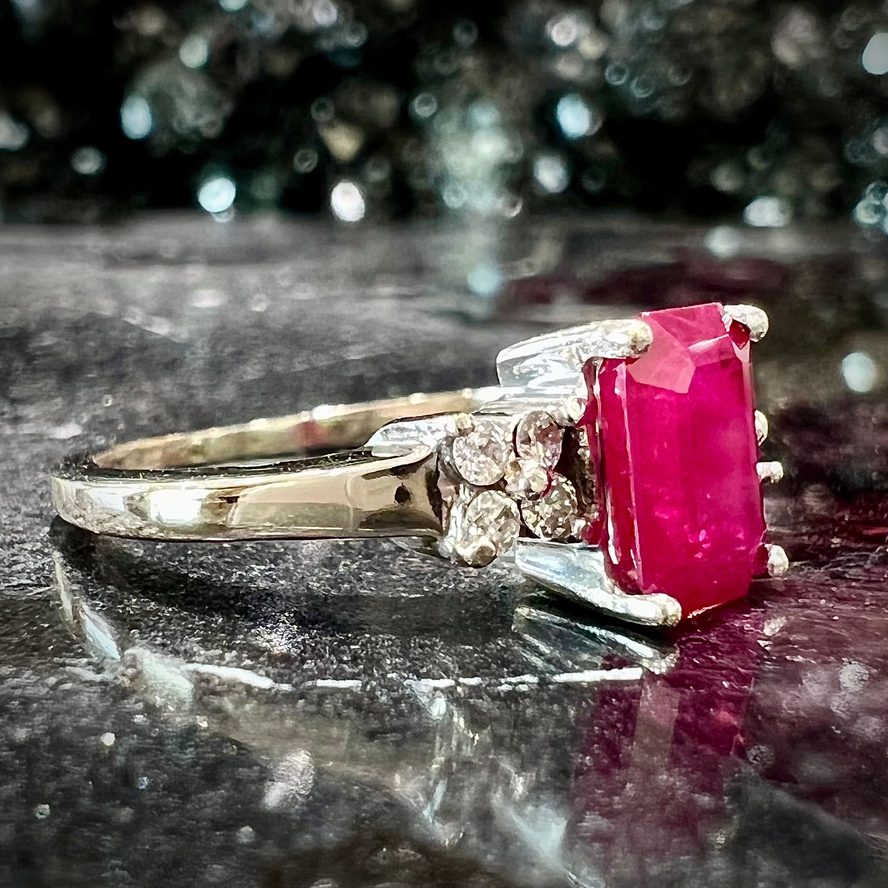 A white gold ring with an emerald cut red synthetic ruby set between four round cut diamonds on each side.