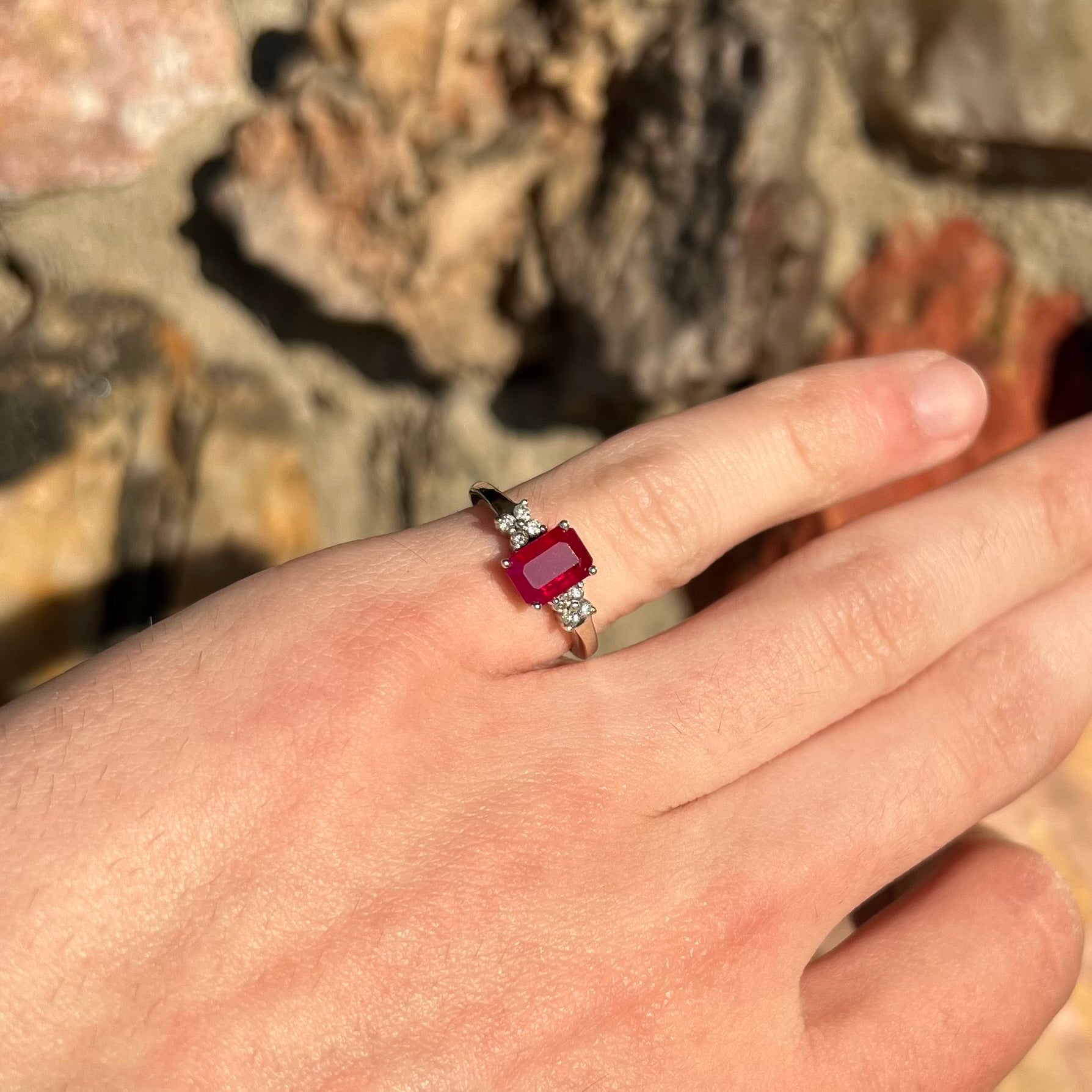 A white gold ring with an emerald cut red synthetic ruby set between four round cut diamonds on each side.